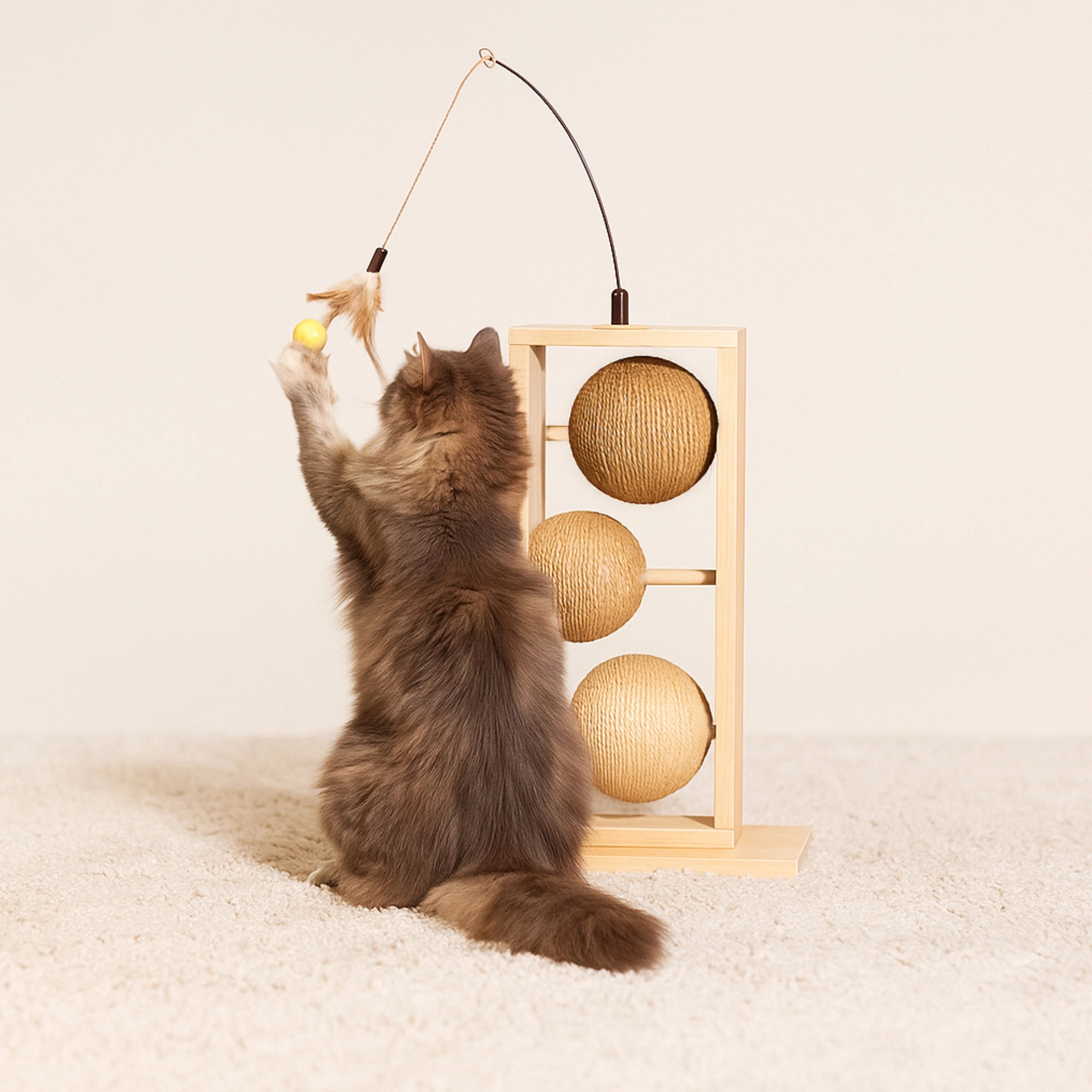 Katze spielt mit Designed by Lotte Kratzbaum Lomax aus hellem Rubberwood mit drei Kratzkugeln aus Jute, Sisal und Seegras sowie integrierter Katzenangel mit Federn – stilvolles, funktionales Kratzmöbel für Katzen.